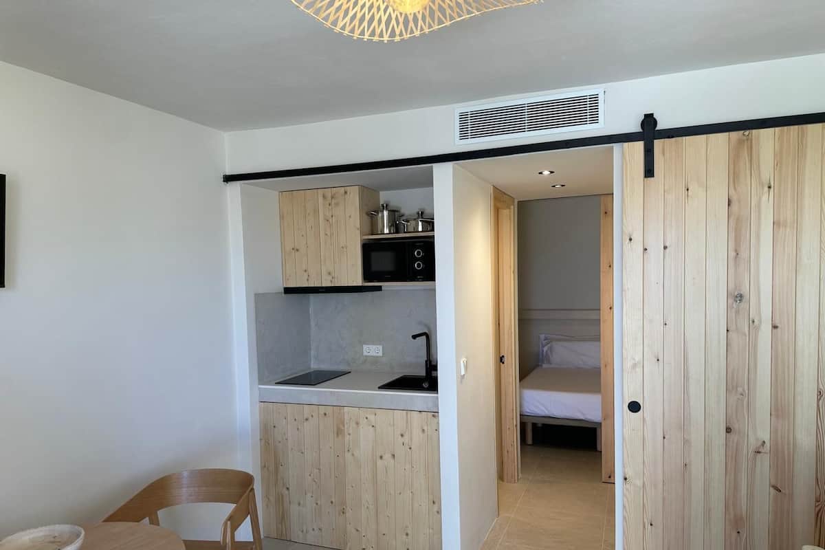 A compact kitchen with wooden cabinets, a microwave, and a sink. There's a dining area with a table and chair, and a bed in the adjacent room.