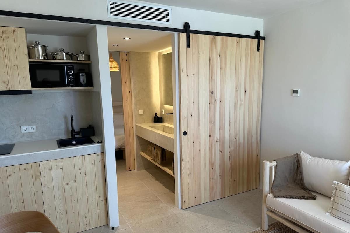 A compact kitchen with a microwave, toaster, and wooden cabinets. A sliding door leads to a bathroom with a mirror and sink. A white sofa is visible in the living area.