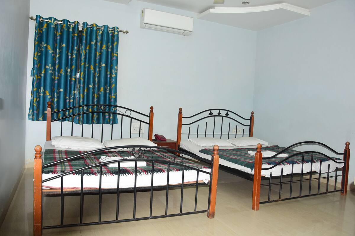 A room with two beds, a window with curtains, an air conditioning unit, and a wooden floor.
