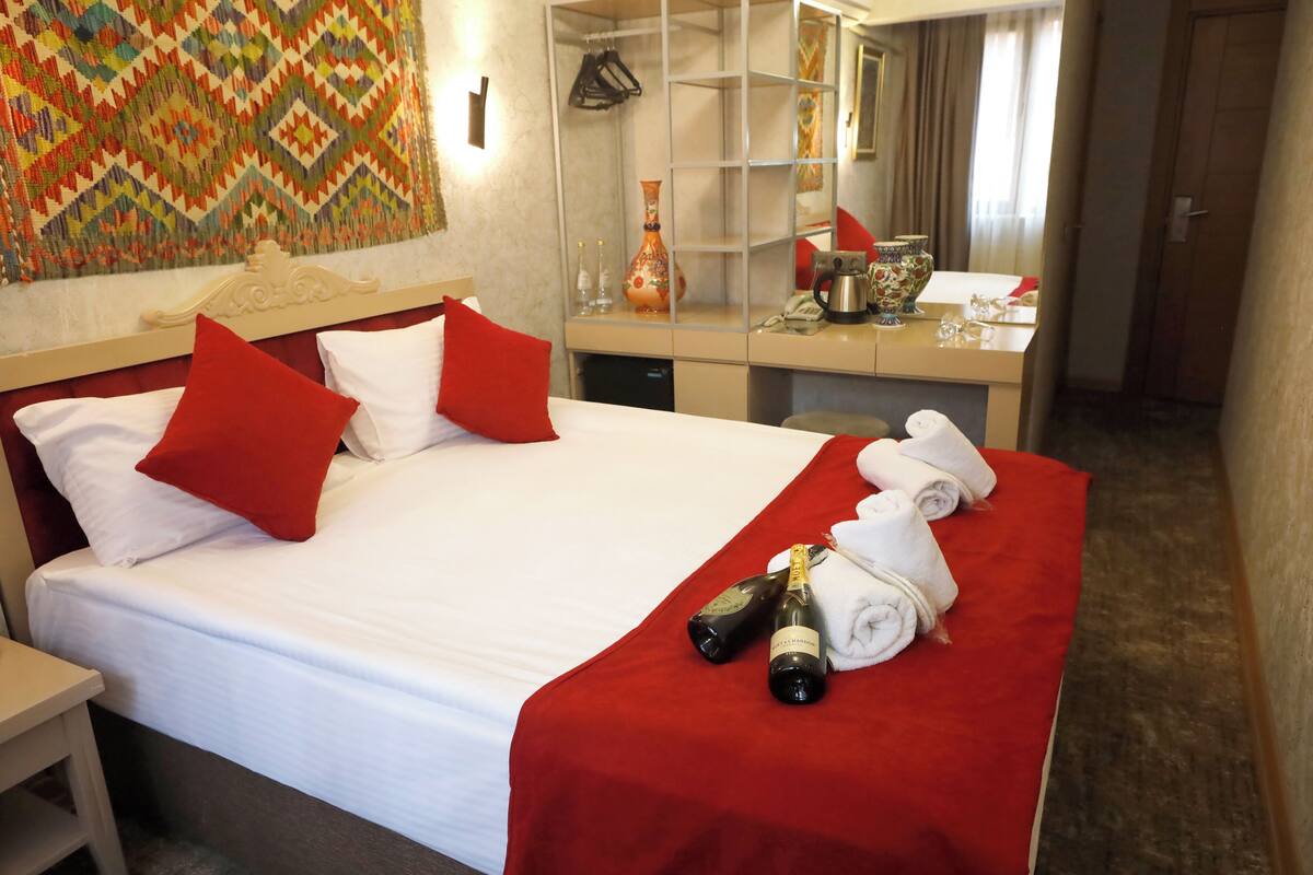 A hotel room with a bed, red pillows, a red bedspread, a minibar, a mirror, and a decorative wall hanging.