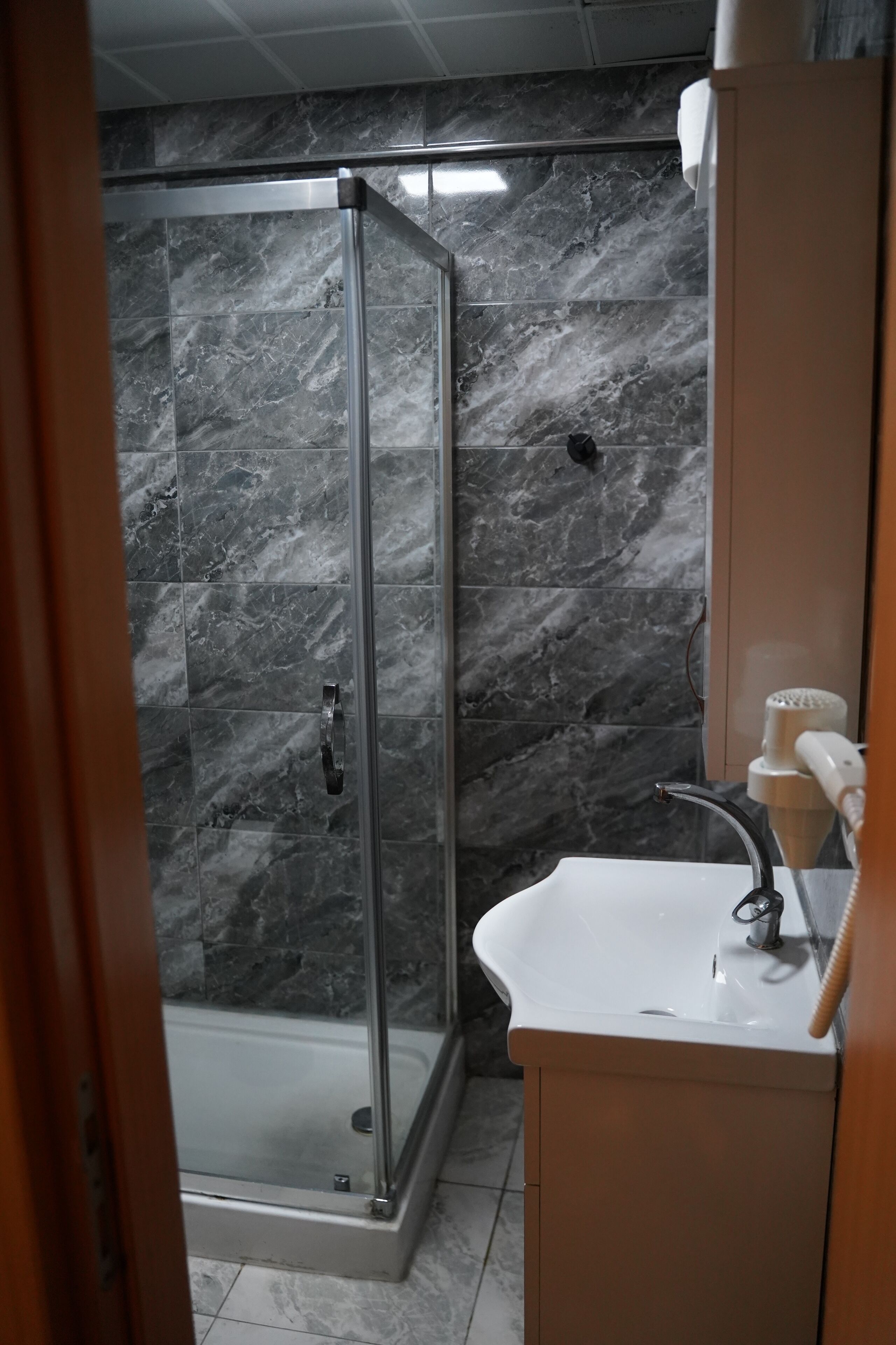 Un baño con ducha envuelta en vidrio, paredes de mármol, un lavamanos blanco y un armario de madera.