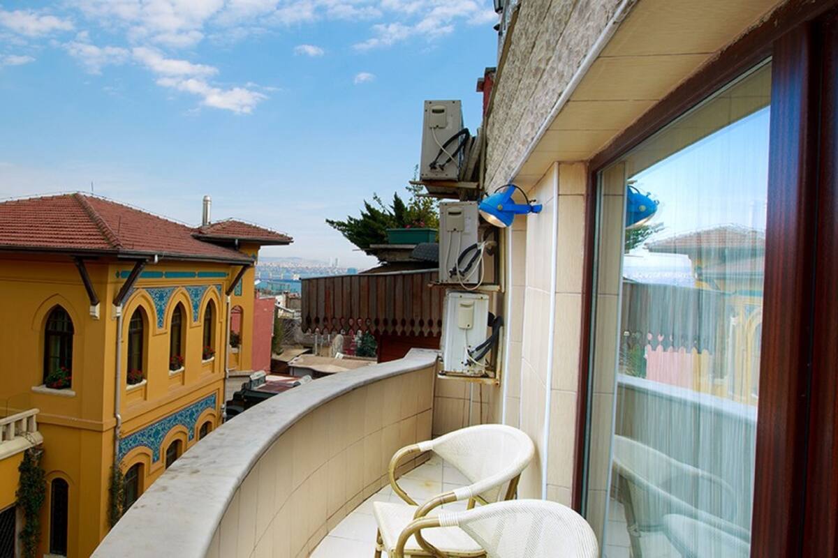 A balcony with a view of a yellow building with blue accents, a white wicker chair, and a large window.
