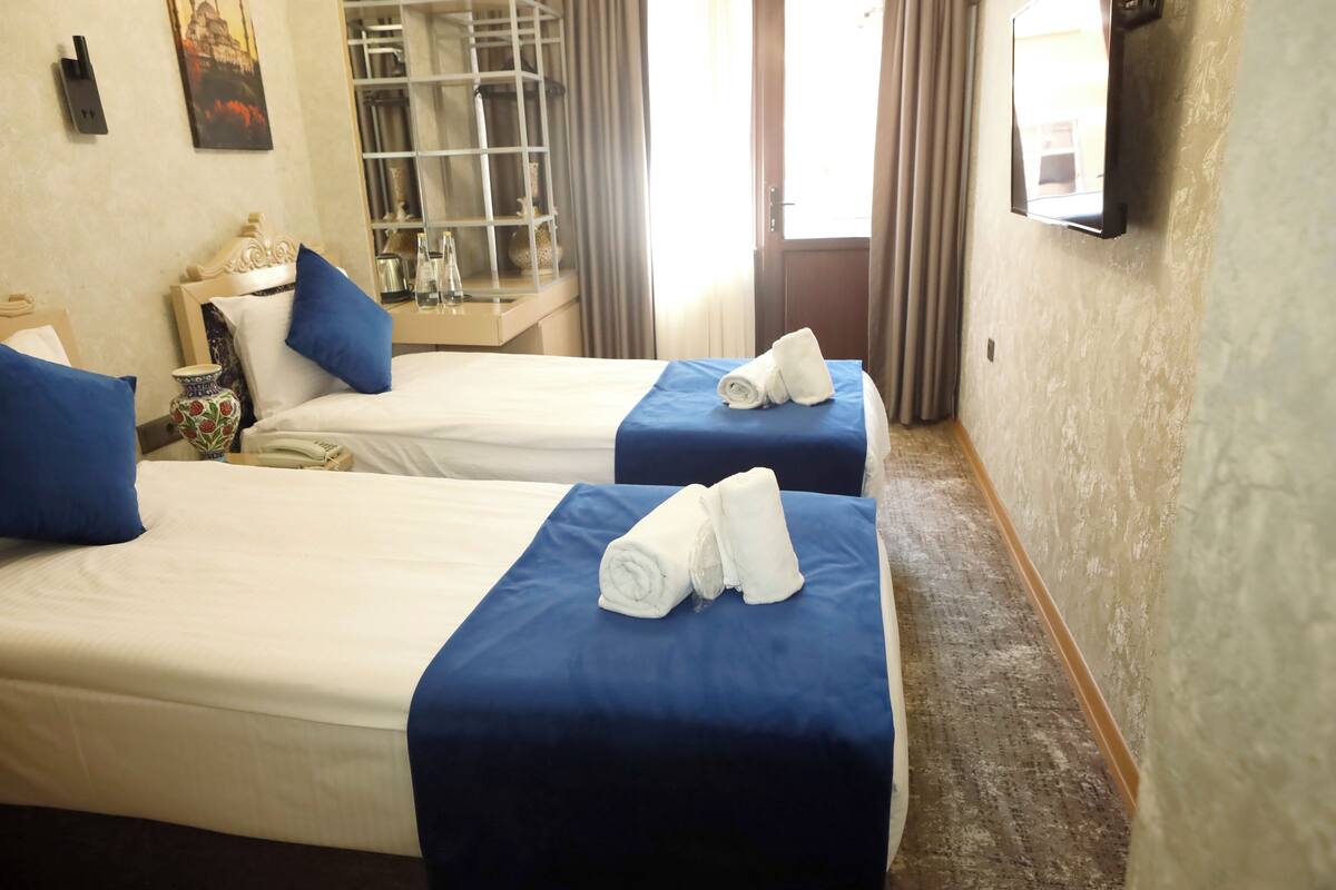 A hotel room with two beds, each with a blue bedspread and white linens, a small table with a vase, and a mirror on the wall.