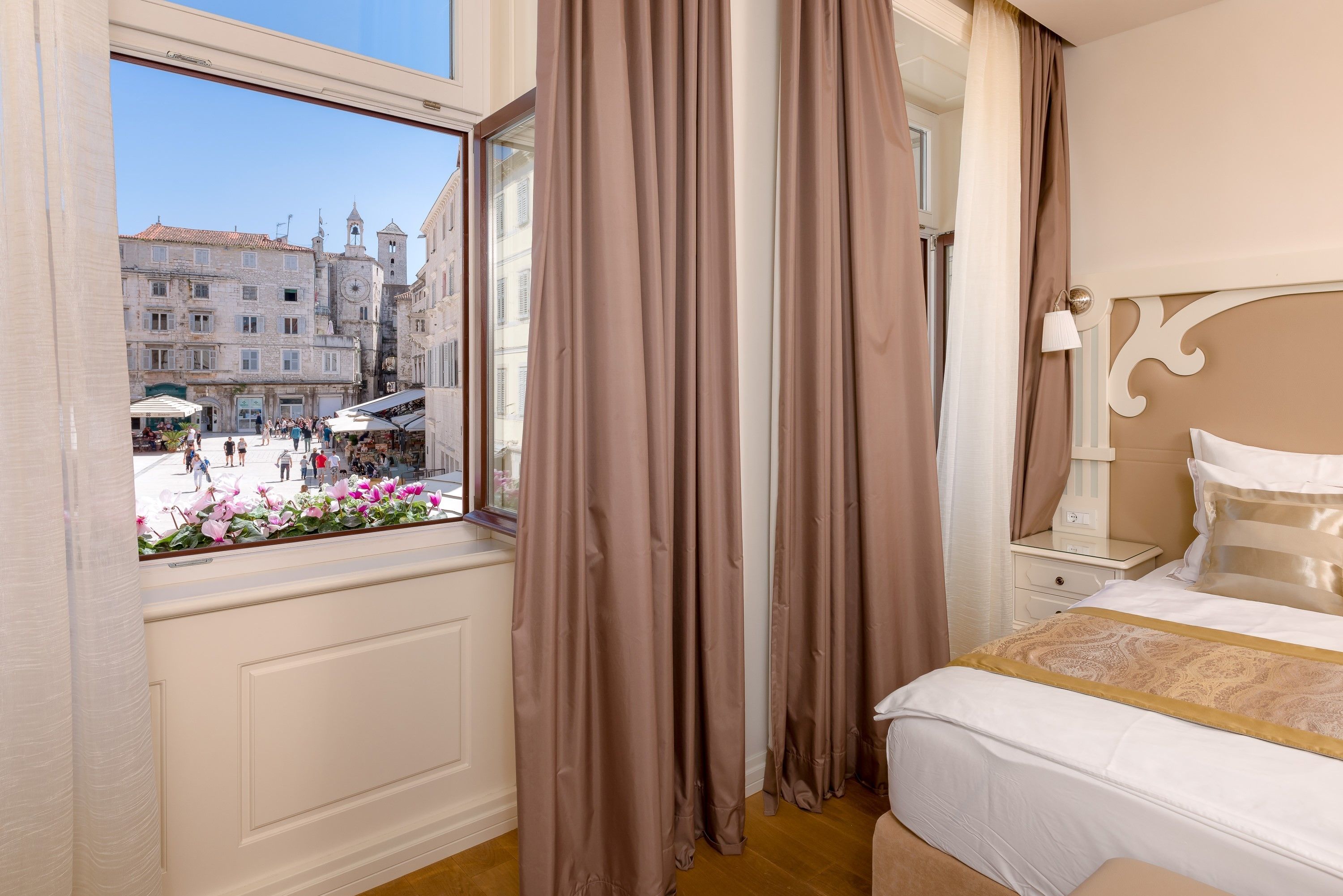 A hotel room with a large window overlooking a public square with buildings and people.