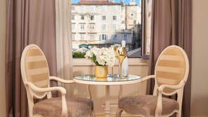 A room with two upholstered chairs and a round glass table, a vase with flowers, and a bottle. A window with curtains reveals a building with a clock tower in the background. - Piazza Heritage Hotel (Split)