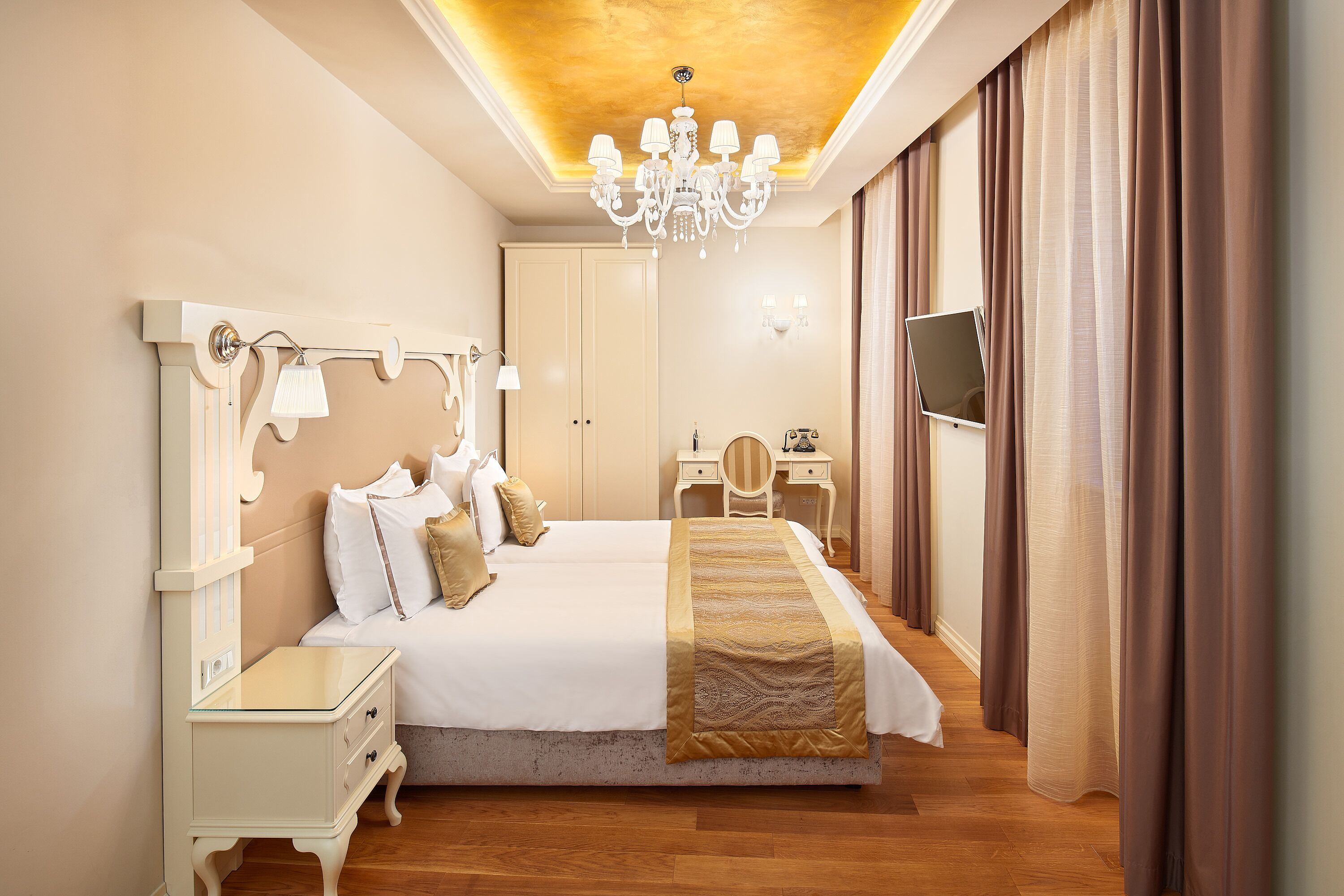 A hotel room with a large bed, bedside tables, a desk, a chair, a vanity with a mirror, and a television on the wall.