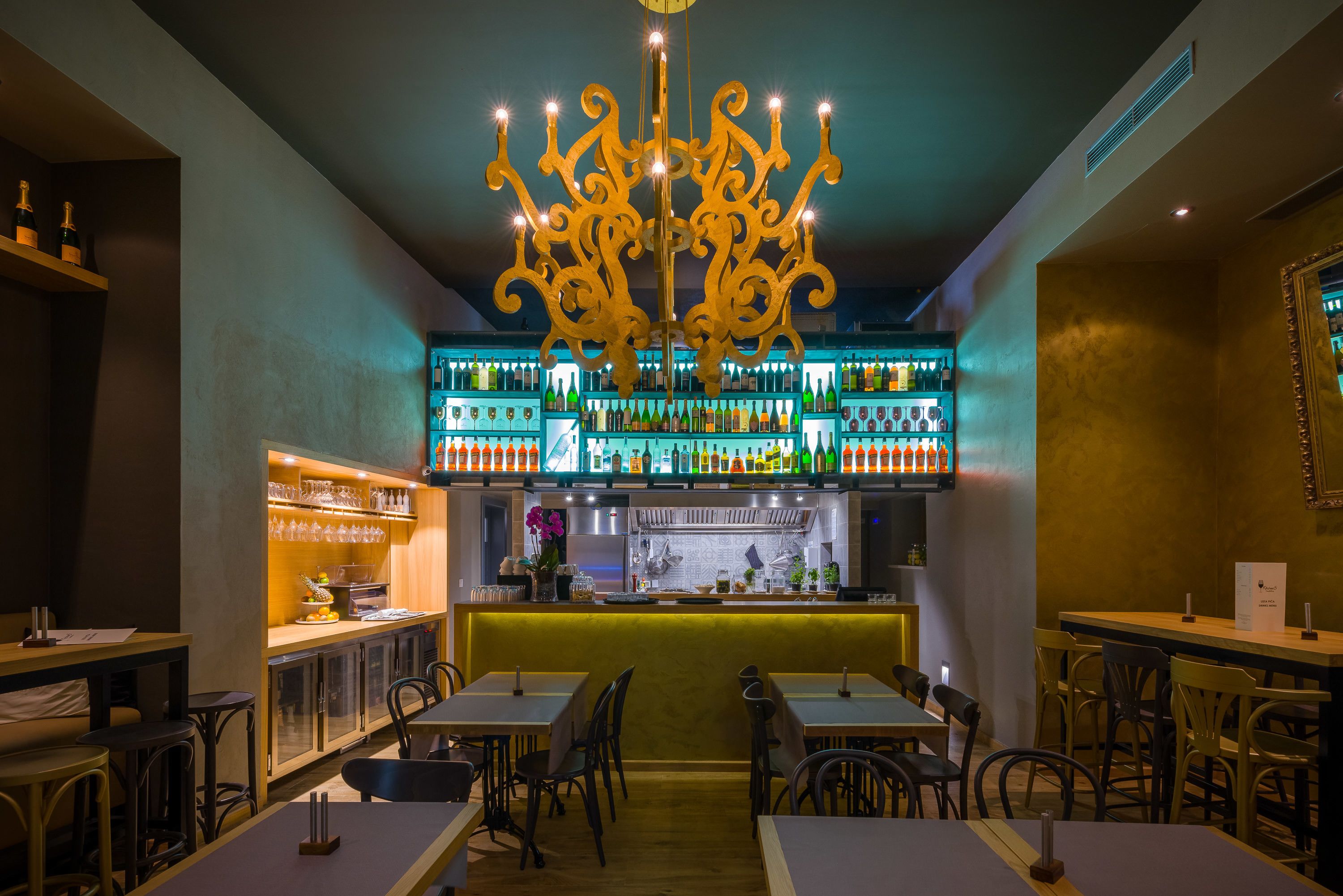 Un espace bar agrémenté d’un lustre central, d’étagères remplies de bouteilles et d’un comptoir équipé d’ustensiles de bar.