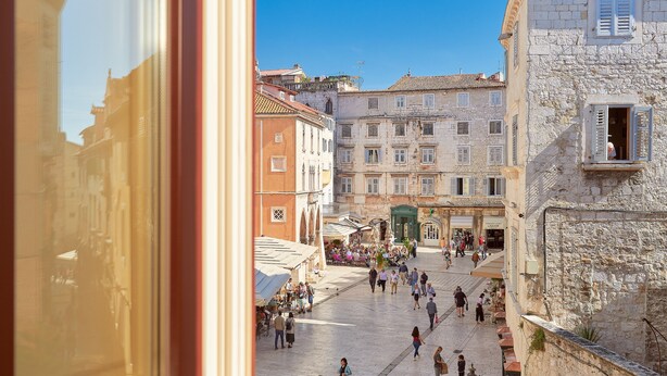 A bustling square with historic buildings, a central open space, and a view through a window. - Piazza Heritage Hotel (Split)