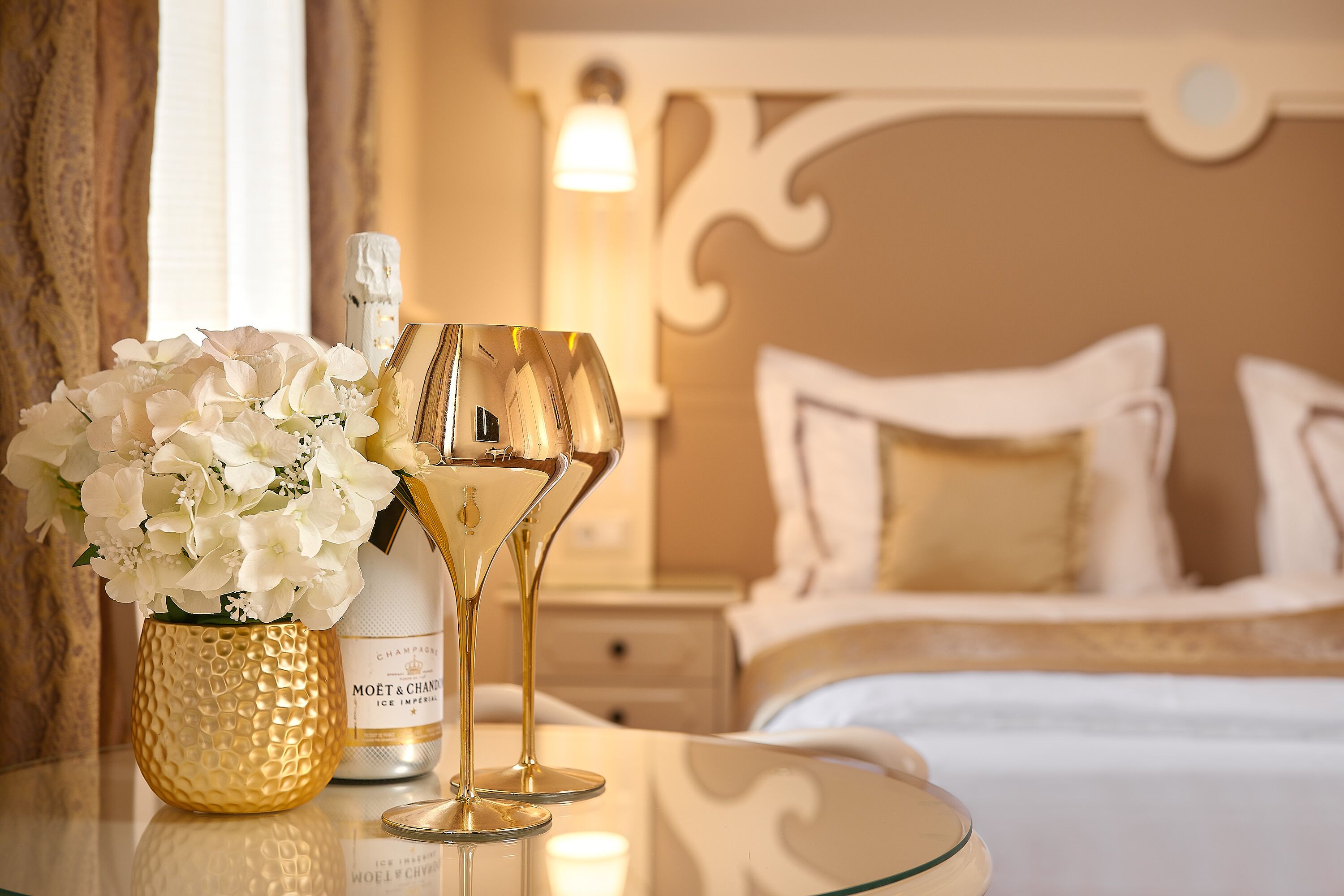 Una cama con ropa blanca, una mesita de noche con una botella de champán y dos copas, y un jarrón con flores blancas.