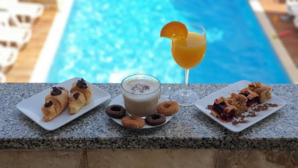 Una mesa con cruasanes, donas, un batido y un cóctel junto a una piscina.
