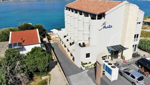 A hotel with a white facade, a red roof, and a view of the sea and mountains. - Hotel Plaža (Pag)