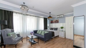 A modern living room with a sofa, armchair, and a small table. There is a bed with a white headboard and a nightstand with a lamp. A chandelier hangs from the ceiling, and a painting is on the wall. - Lavanta Hotel (Istanbul)