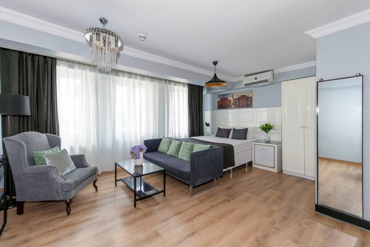 A modern living room with a sofa, armchair, and a small table. There is a bed with a white headboard and a nightstand with a lamp. A chandelier hangs from the ceiling, and a painting is on the wall.