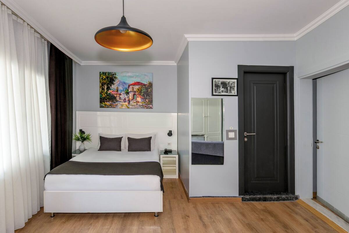 A modern bedroom with a large bed, a bedside table with a lamp, a painting on the wall, a door, and a mirror.