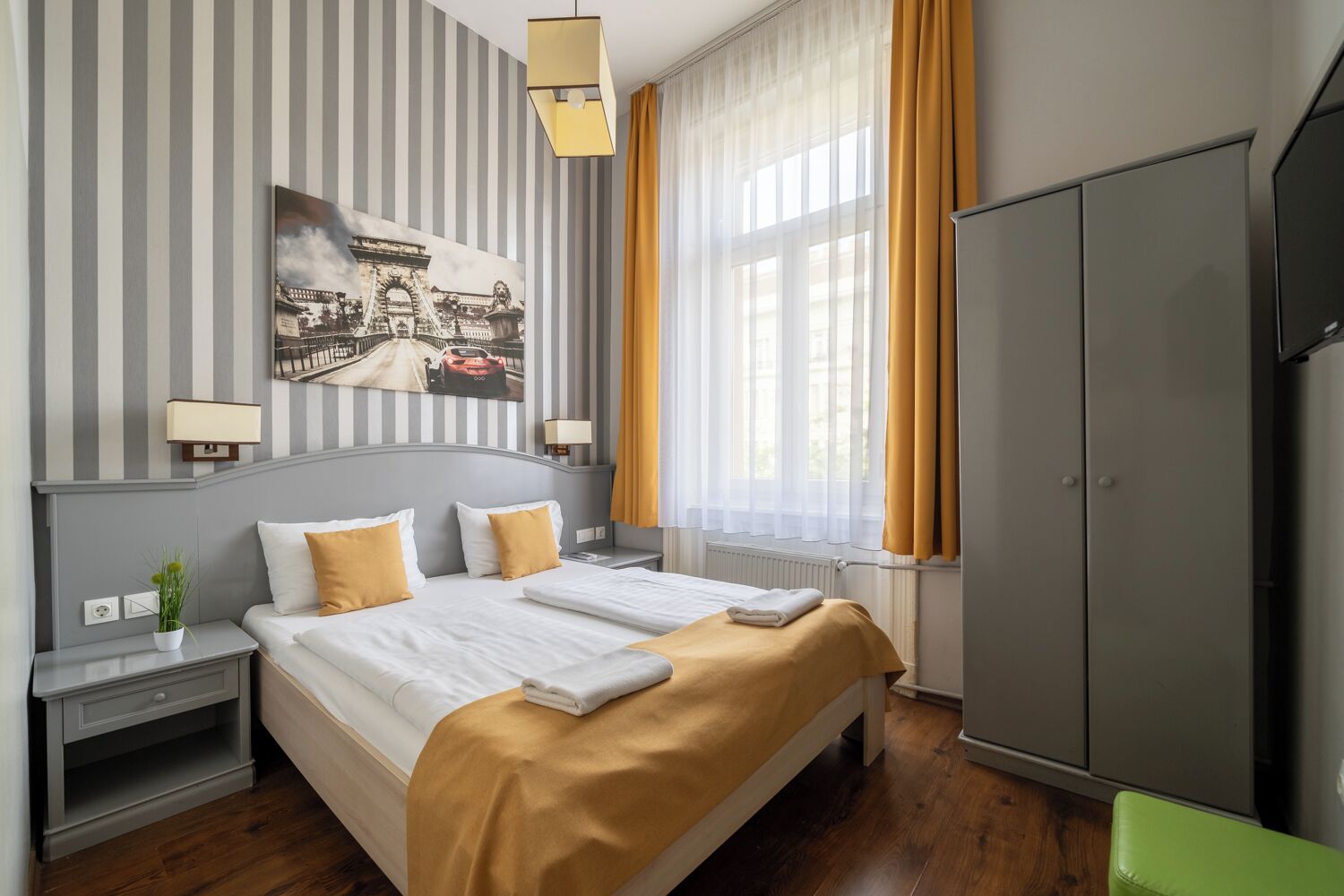 A hotel room with a large bed, two bedside tables, a nightstand, a wardrobe, and a window with curtains.