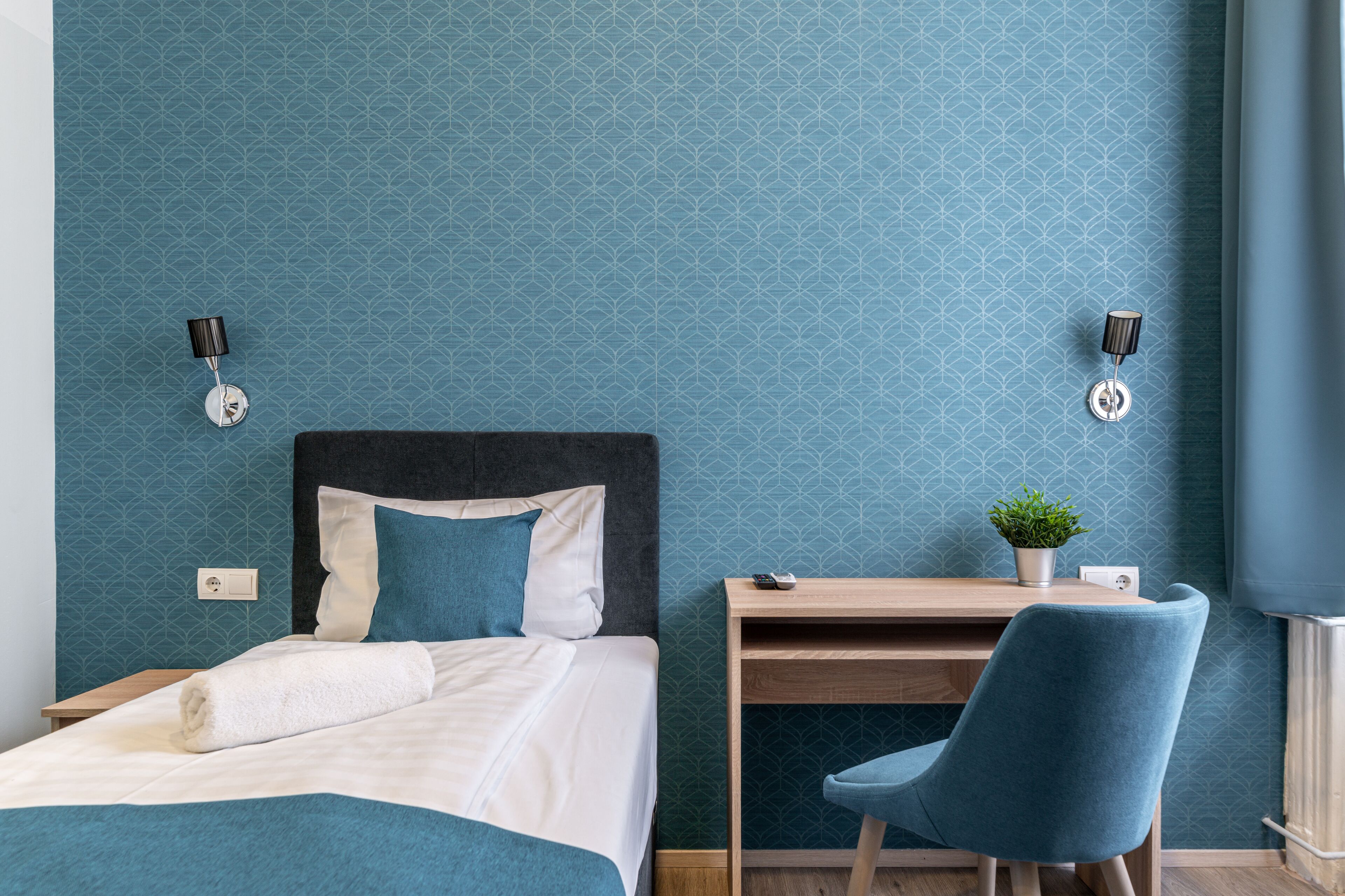 A hotel room with a bed, a desk, a chair, and a wall-mounted lamp.