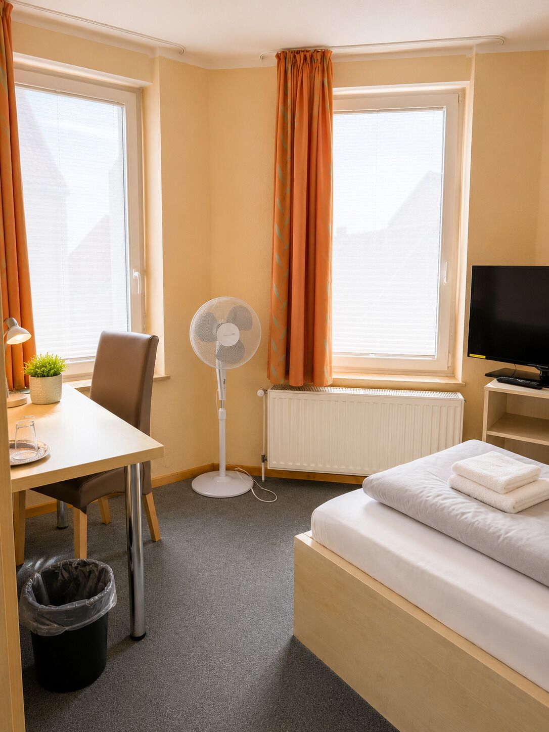A hotel room with a bed, a desk with a chair, a standing fan, a TV, and a window with curtains.