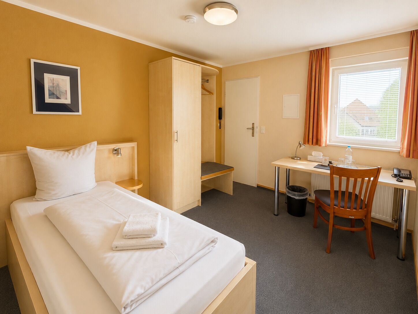 Une chambre d’hôtel avec un lit, un bureau, une chaise, une armoire, un tableau au mur et une fenêtre avec des rideaux.