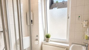 A modern bathroom with a glass shower enclosure, a toilet, a sink, and a window with a view of a building. - Hotel Select (Moenchengladbach)