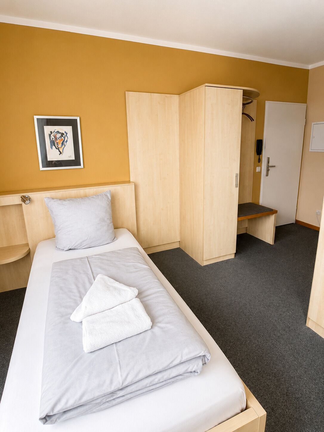 A single bed with a white mattress and grey sheets, a white pillow, a wooden wardrobe, a small bench, and a door with a phone.