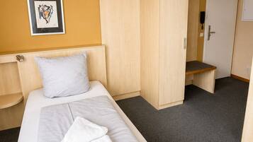 A single bed with a white mattress and grey sheets, a white pillow, a wooden wardrobe, a small bench, and a door with a phone.