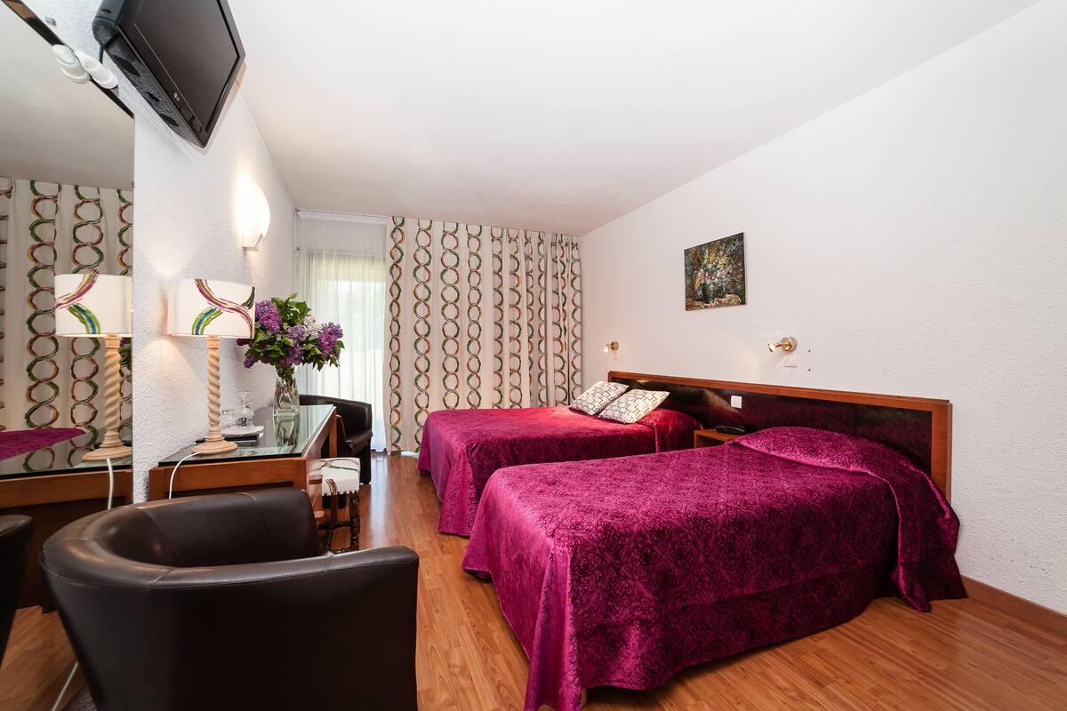 A hotel room with two beds, a desk with a chair, a TV mounted on the wall, and a window with patterned curtains.