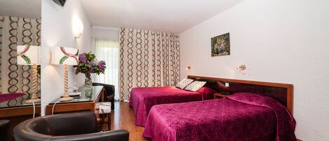 A hotel room with two beds, a desk with a chair, a TV mounted on the wall, and a window with patterned curtains.