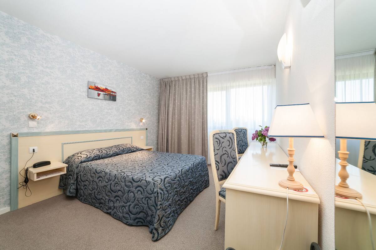 A hotel room with a bed, a desk, a chair, a lamp, a telephone, and a picture on the wall.