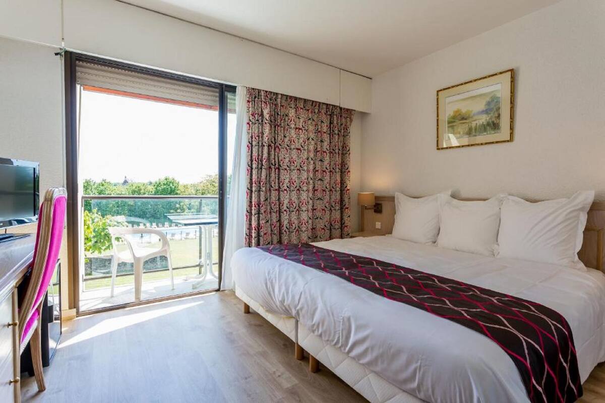 A hotel room with a bed, a desk with a chair, a television, and a balcony with a view of greenery.