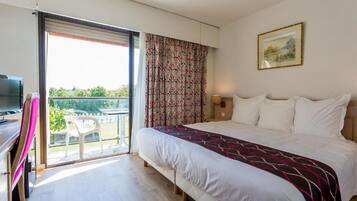 Habitación de hotel con cama, escritorio con silla, televisor y balcón con vista a follaje.
