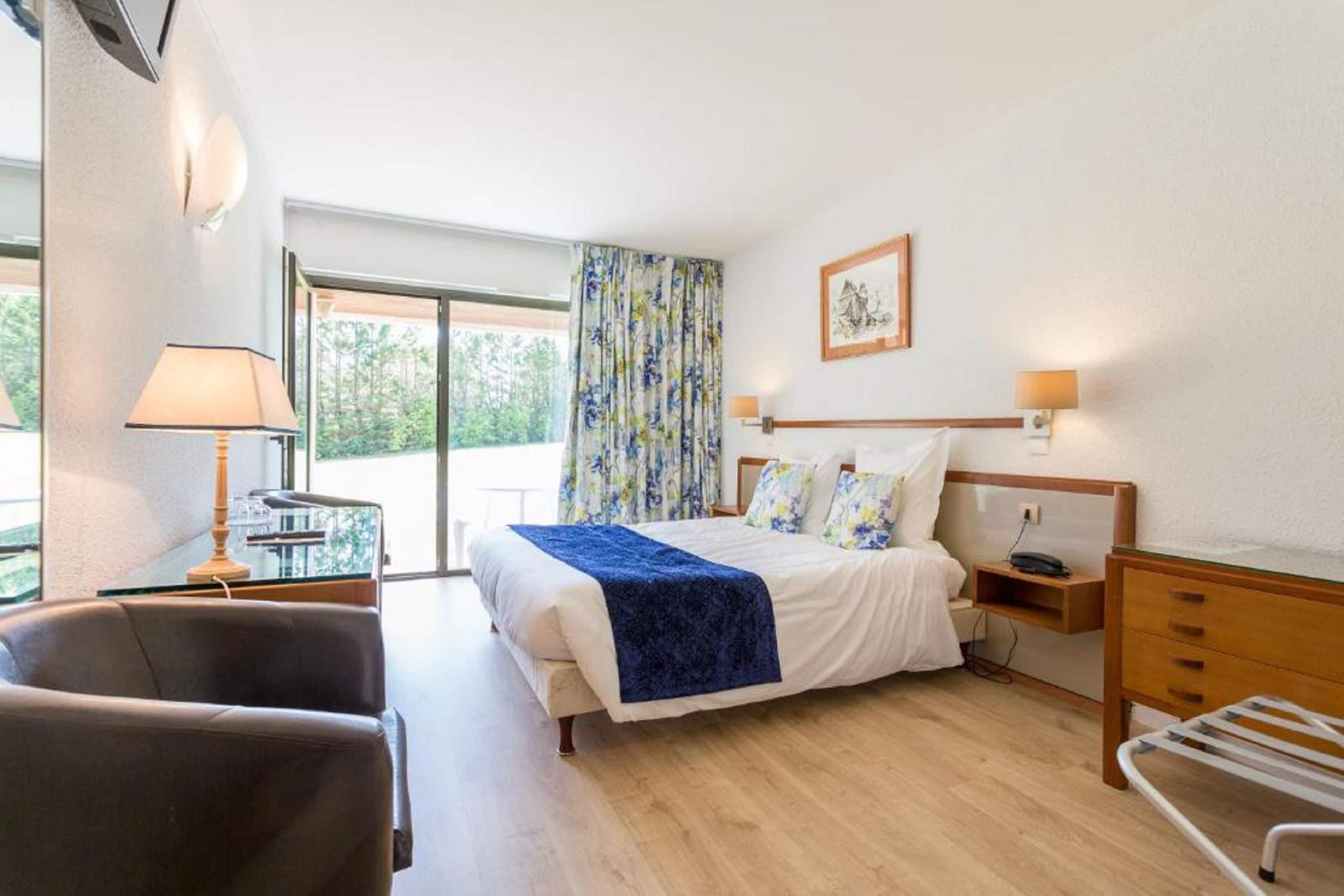 Un dormitorio con cama, mesita de noche, lámpara, silla y vistas al exterior.