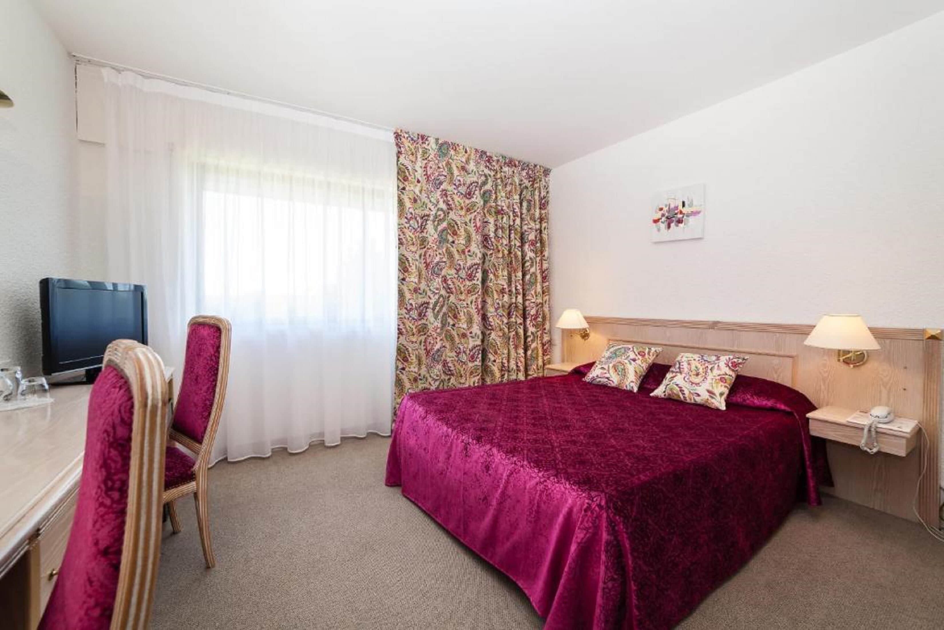 Une chambre d’hôtel comprenant un lit, un bureau avec une chaise, une télévision et une fenêtre avec des rideaux.