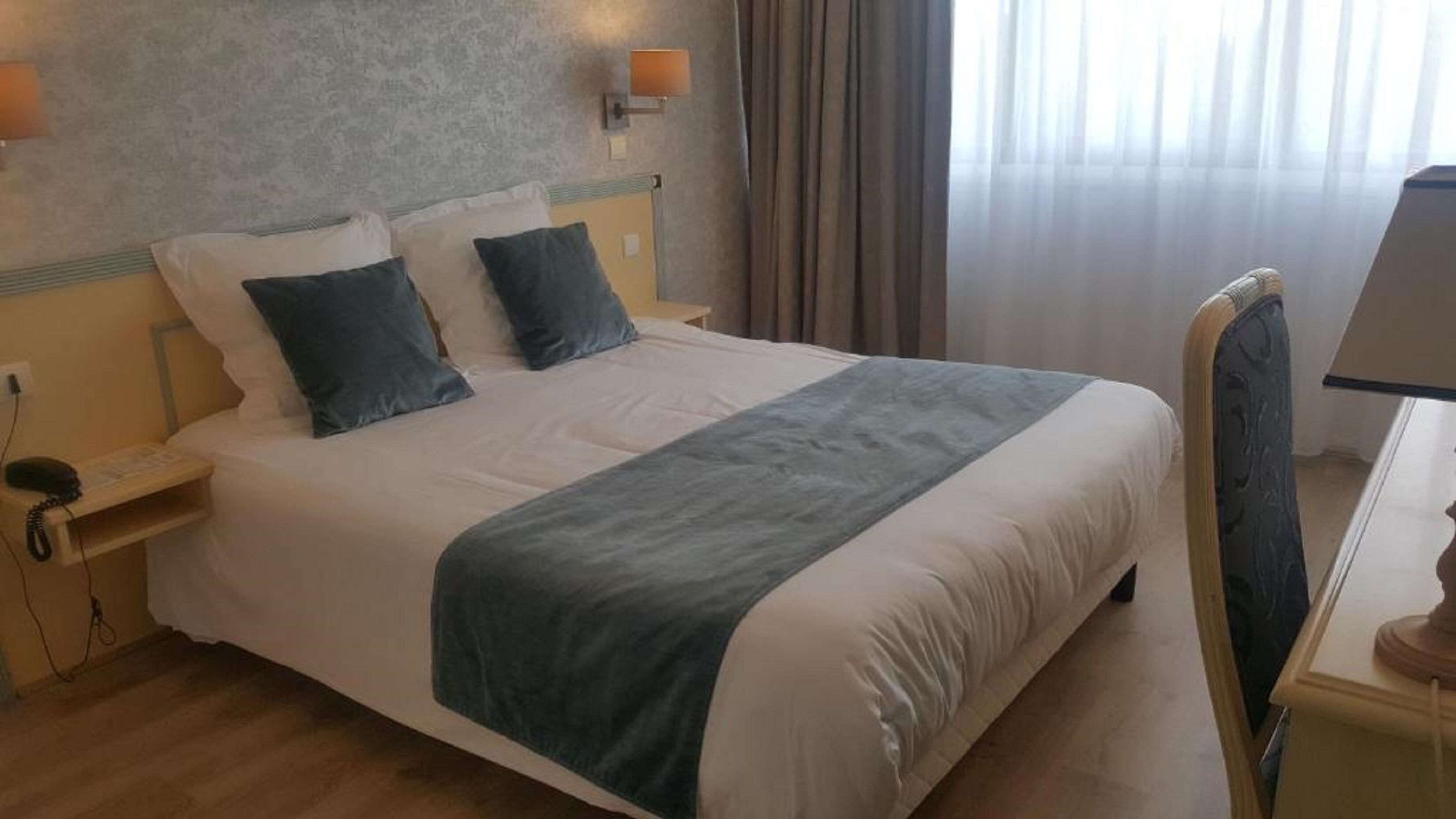 Quarto de hotel com uma cama grande, duas mesas de cabeceira, uma escrivaninha com abajur e uma janela com cortinas.