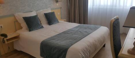 A hotel room with a large bed, two bedside tables, a desk with a lamp, and a window with curtains.