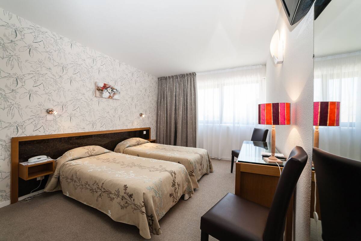 A hotel room with two beds, a desk, a chair, a TV, and a window with curtains.
