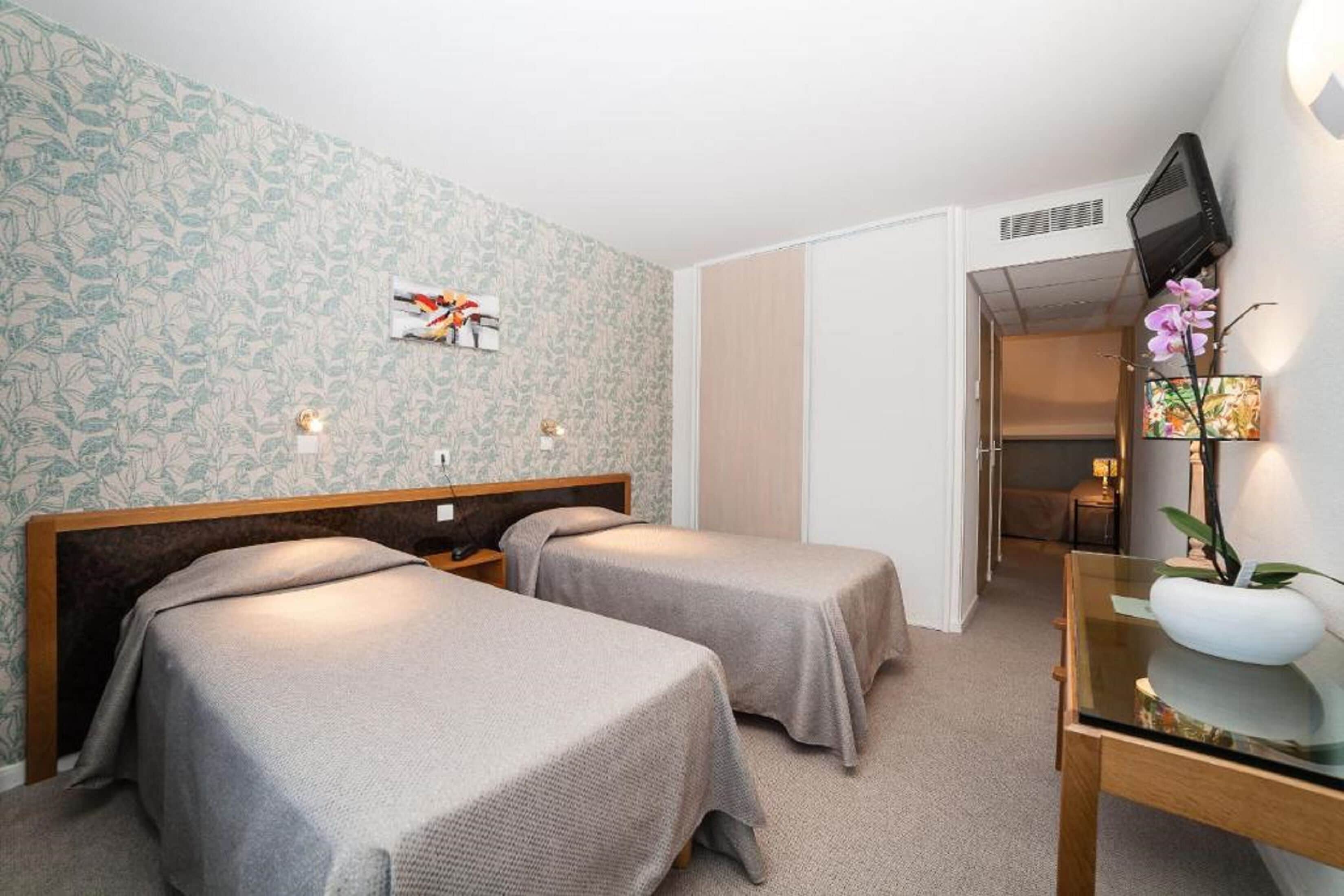 Une chambre d’hôtel avec deux lits simples, une tête de lit en bois, une petite table avec un vase et un téléviseur fixé au mur.