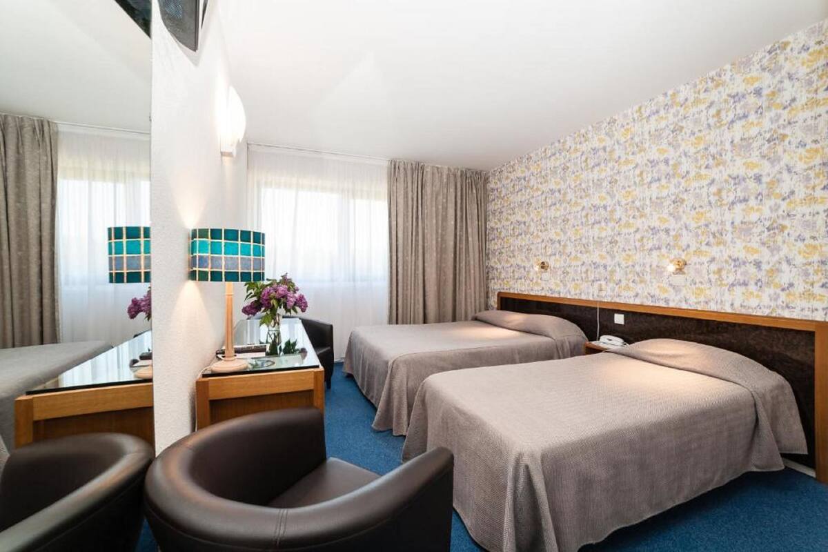 A hotel room with two beds, a desk with a lamp, a chair, and a view of the outdoors.