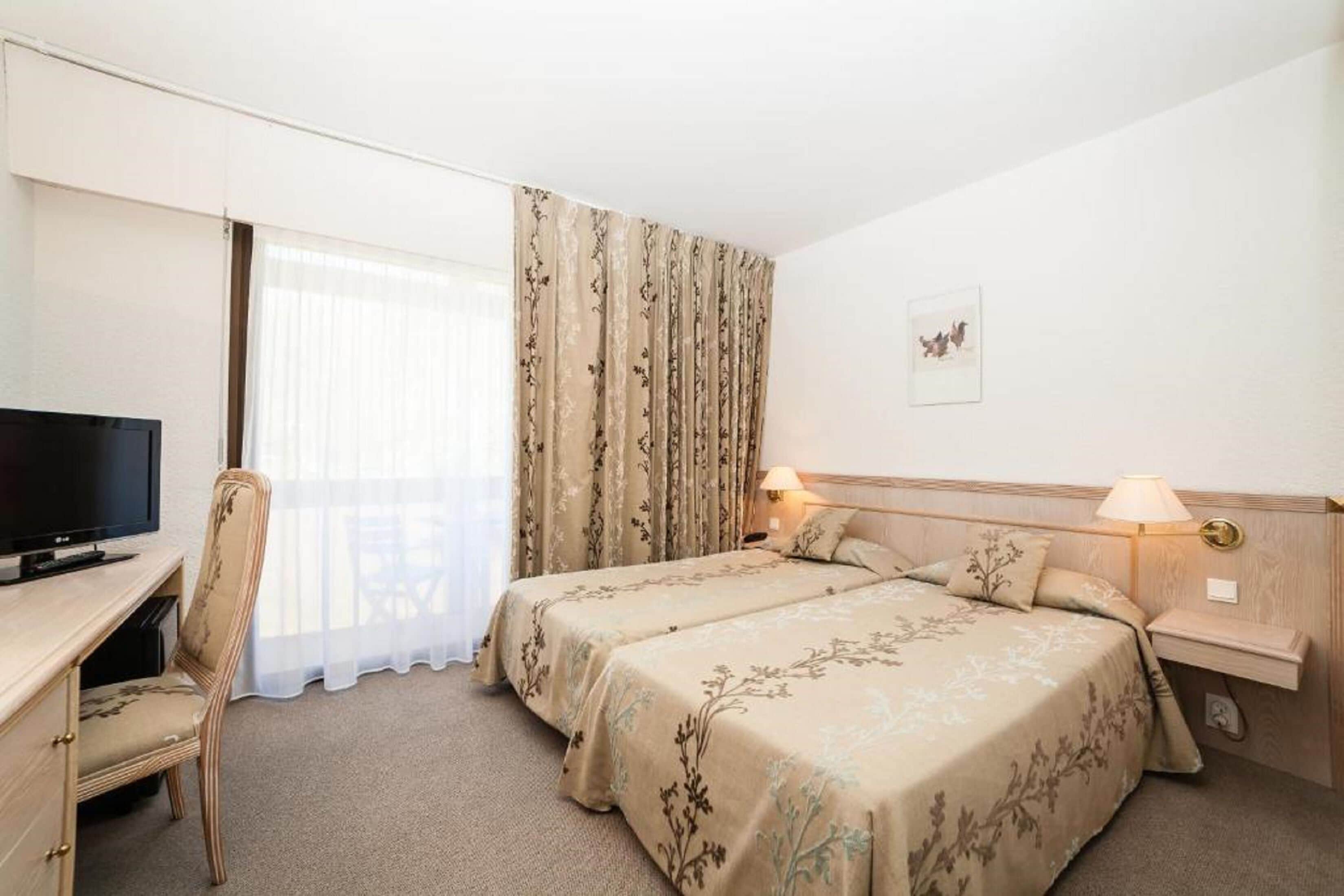 A hotel room with two beds, a desk with a TV, a chair, and a window with curtains.