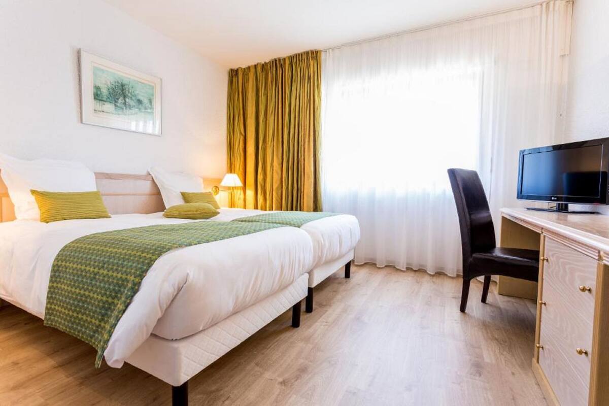 A hotel room with two beds, a desk, a TV, and a window with curtains.