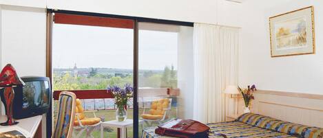 Ein Hotelzimmer mit Bett, Schreibtisch mit Fernseher, Stuhl, Tisch mit Blumenvase und Blick ins Freie.