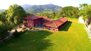 A red building with a tiled roof, surrounded by a well-maintained lawn and trees, set against a backdrop of hills. - Coviella Hotel Rural (Cangas de Onis)