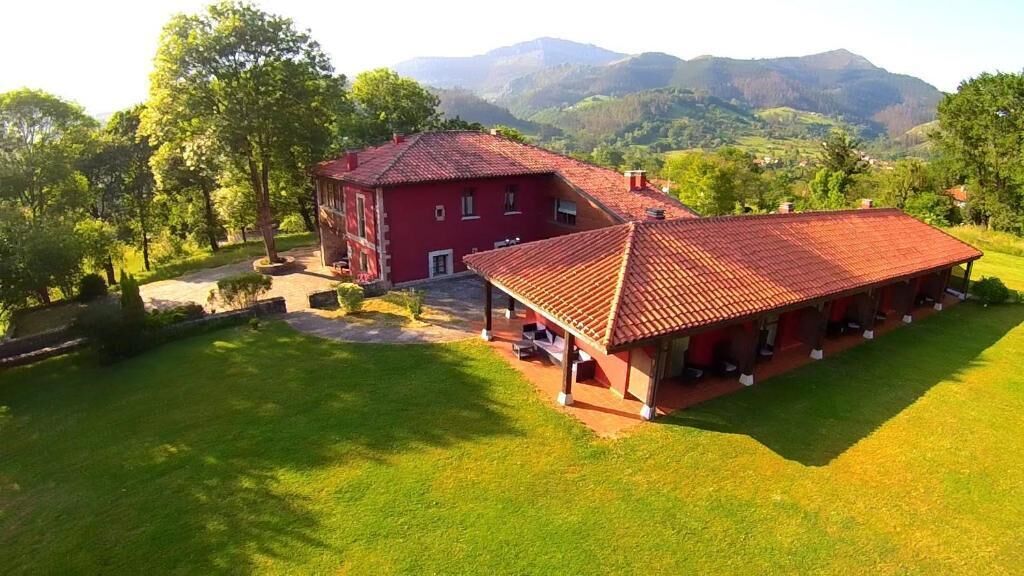 Un edificio de techo rojo con un área de estar al aire libre cubierta, rodeado de exuberante vegetación y montañas al fondo.