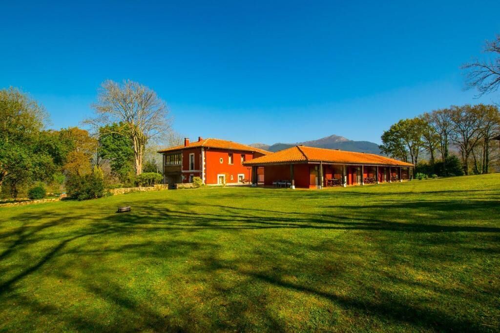 A large, red building with a spacious lawn in front, surrounded by trees and mountains in the background.