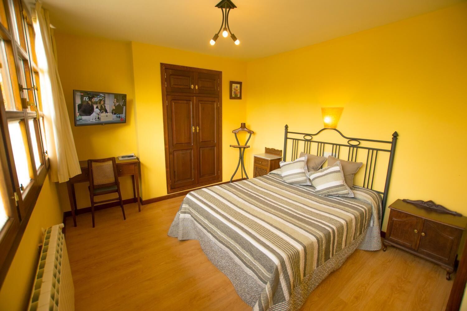 Un dormitorio con cama, un armario de madera, una mesita de noche, una silla, un escritorio y una ventana con cortinas.