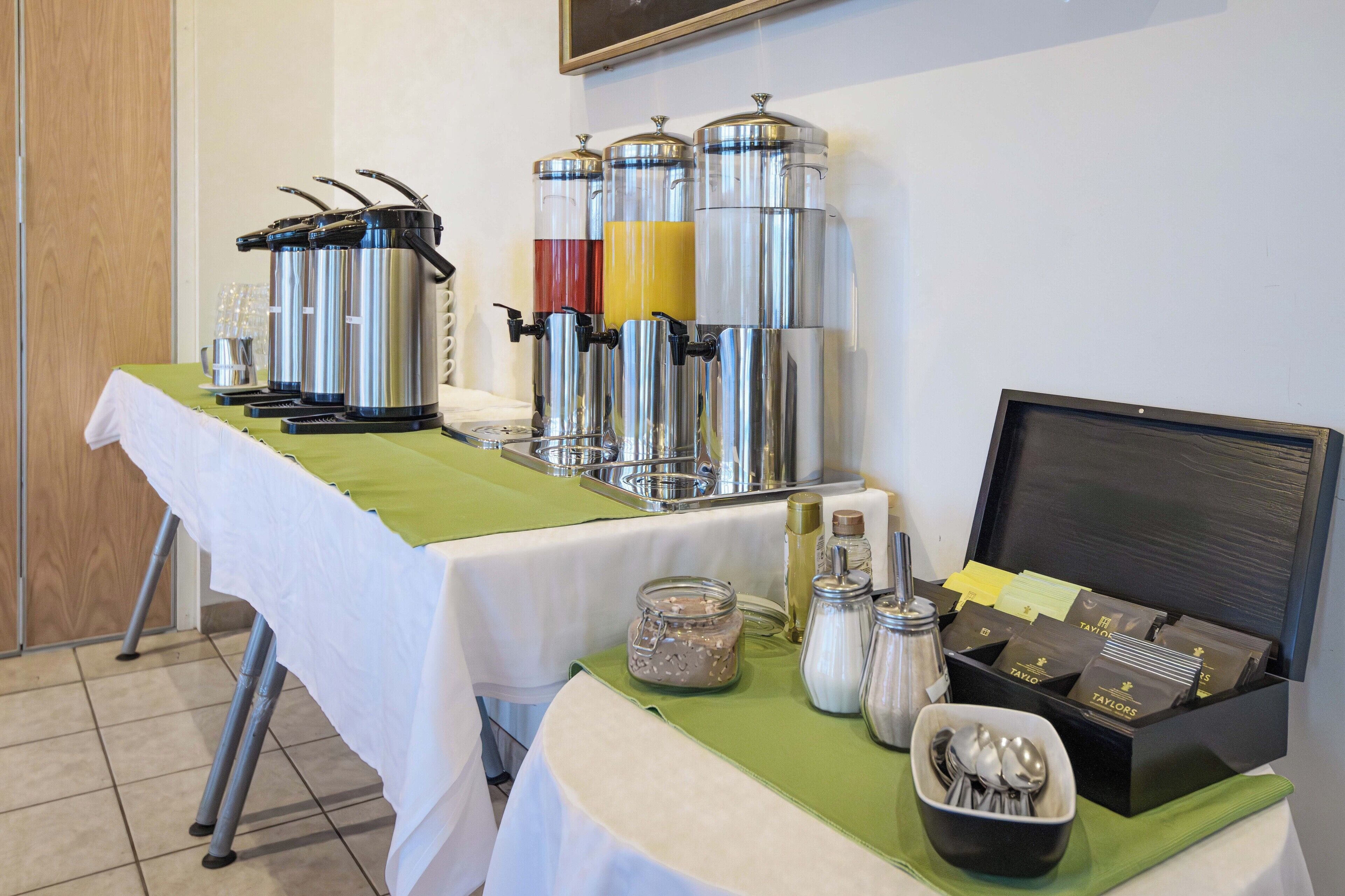 Una mesa de buffet con diferentes dispensadores de comida y bebida, una máquina de café y una caja de bolsitas de té.