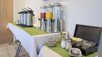 Una mesa de buffet con diferentes dispensadores de comida y bebida, una máquina de café y una caja de bolsitas de té.