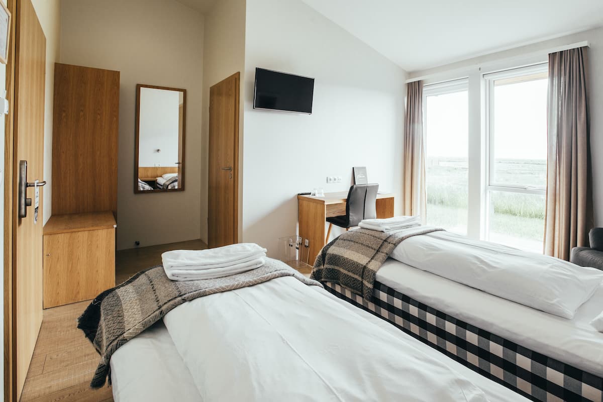 Habitación de hotel con dos camas, un escritorio, un televisor y un ventanal grande con cortinas.