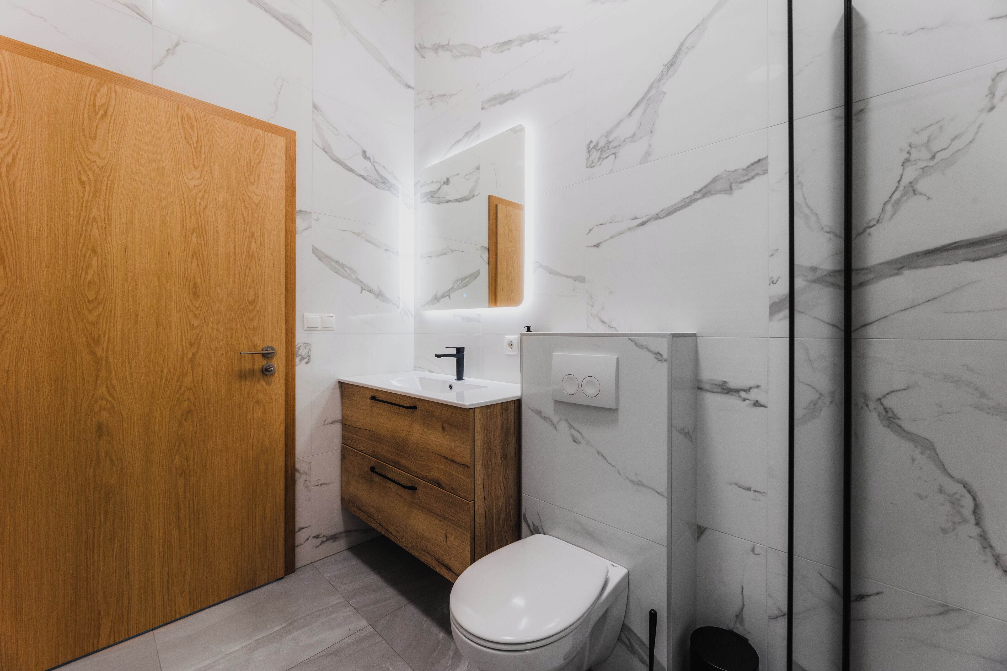 A modern bathroom with a wooden door, marble walls, a white sink, and a toilet.
