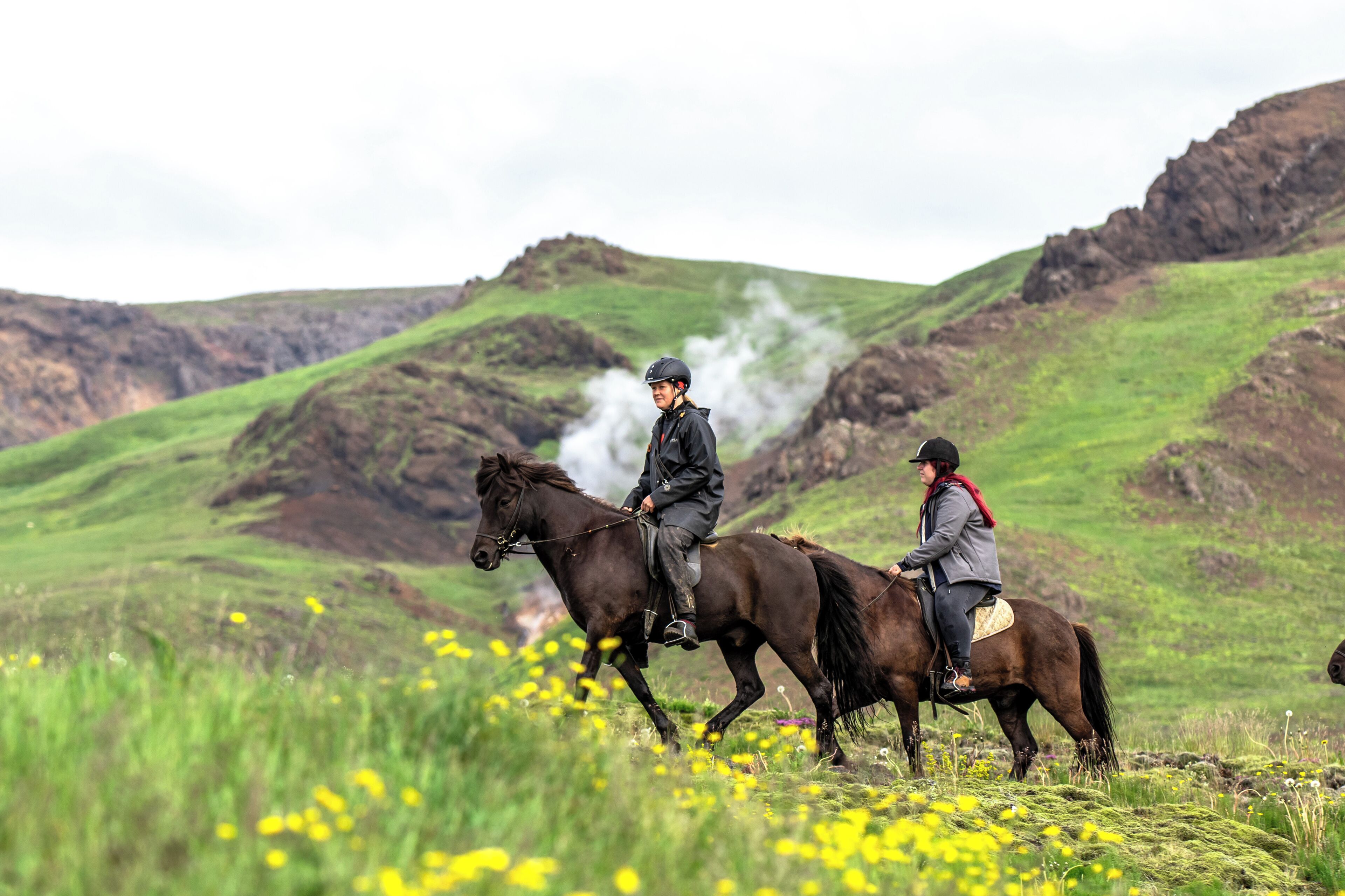 Deux personnes à cheval, évoluant sur un champ herbeux, avec une chaîne de montagnes en arrière-plan.