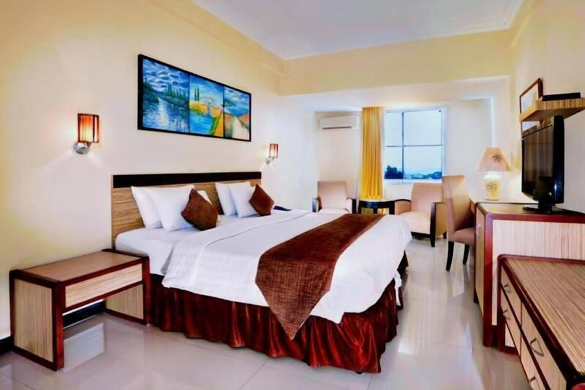 Una habitación de hotel con cama, mesitas de noche, escritorio, silla, televisor y un cuadro en la pared.